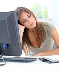 A young woman, facing the viewer, sitting behind a desk with a computer monitor, looking at a book.