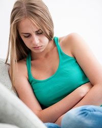 A young woman facing the viewer, sitting on a sofa looking downcast, with her arms folded across her abdomen, looking like she is physically uncomfortable.