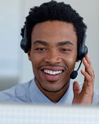 A smiling young man facing the viewer wearing headphones that have a microphone on a plastic arm that extends from one side to the side of his mouth.