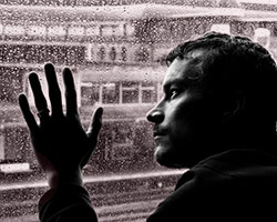 A dreary black and white picture with a man looking at his hand on a window, with the scene outside made up of bleak looking, run-down factories and rail yards.