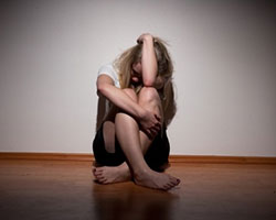 A despondent young woman sits facing the viewer on the floor with her knees pulled up close and her left arm resting on them and obscuring her face.