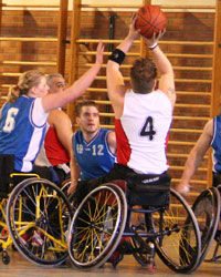A mixed group of 3 wheelchair basketball players in a gym, making a play for a basketball floating in the air, while a fourth person, standing, is looking on.