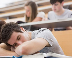 A young man in a classroom with his head lying on his desk with eyes closed.