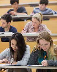 Several students sitting in a classroom, facing the camera, busily taking notes with pen and paper and one on a tablet device. Some likely have disabilities but if so, they are not visible ones.